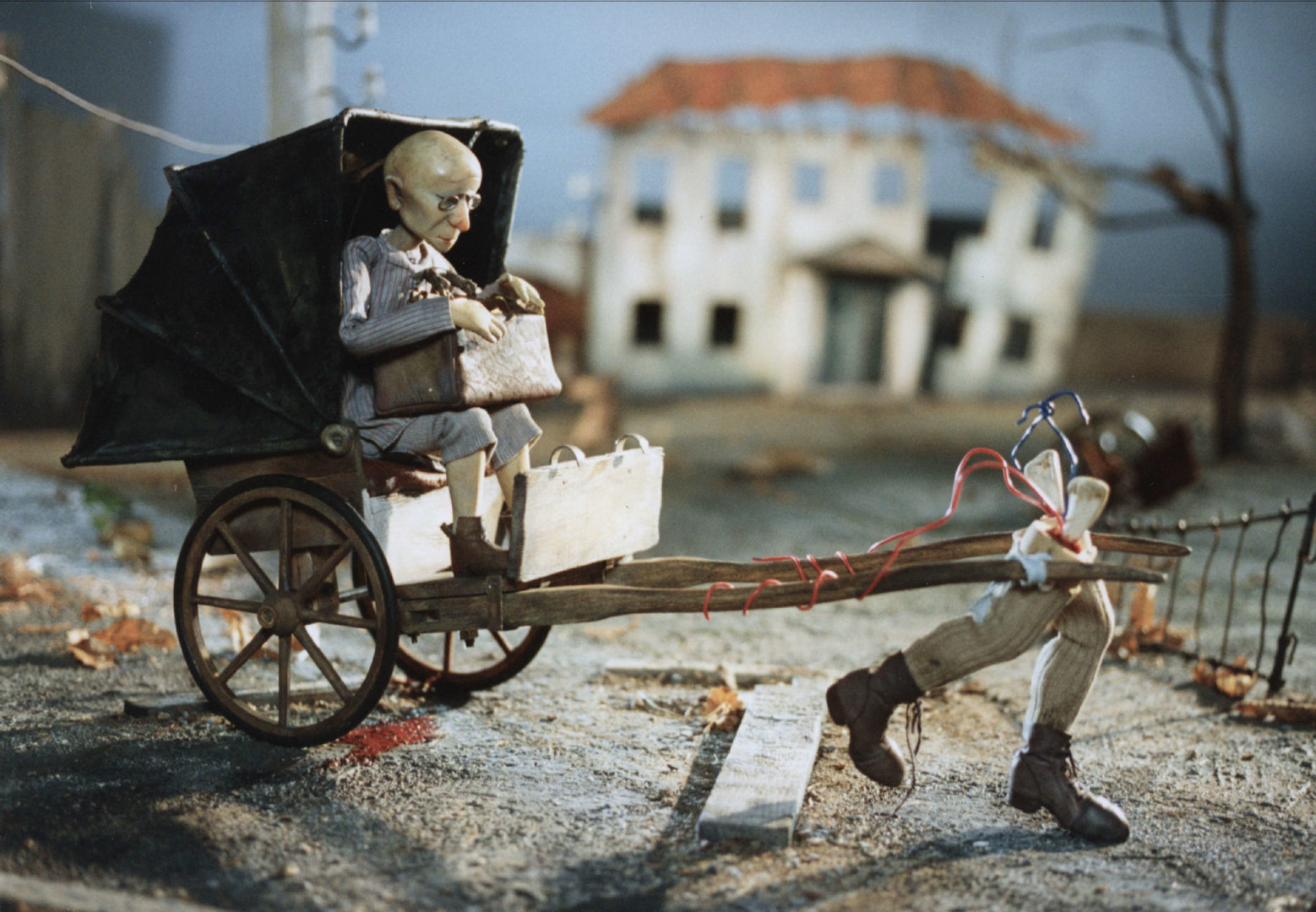 Still picture from The Country Doctor (Maalaislääkäri) from 1996. The picture shows an elderly person travelling in a carriage in grey weather while lower limbs are pulling the carriage.