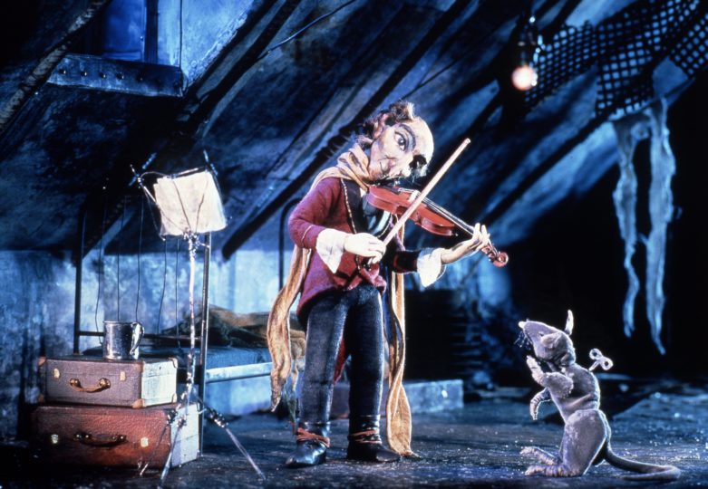 Still picture from the film Rider on the Bucket (Hiilisangolla ratsastaja) from 1992. The picture shows a person playing a violin in a dimly lit room while a rat listens nearby.